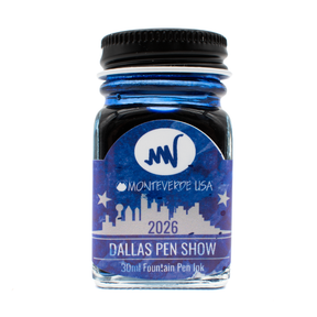 A clear glass fountain pen ink bottle with a black metal cap. The blue Monteverde USA '2026 Dallas Pen Show' label shown over the bottle which reveals a matching royal blue ink underneath shown against a plain white background.