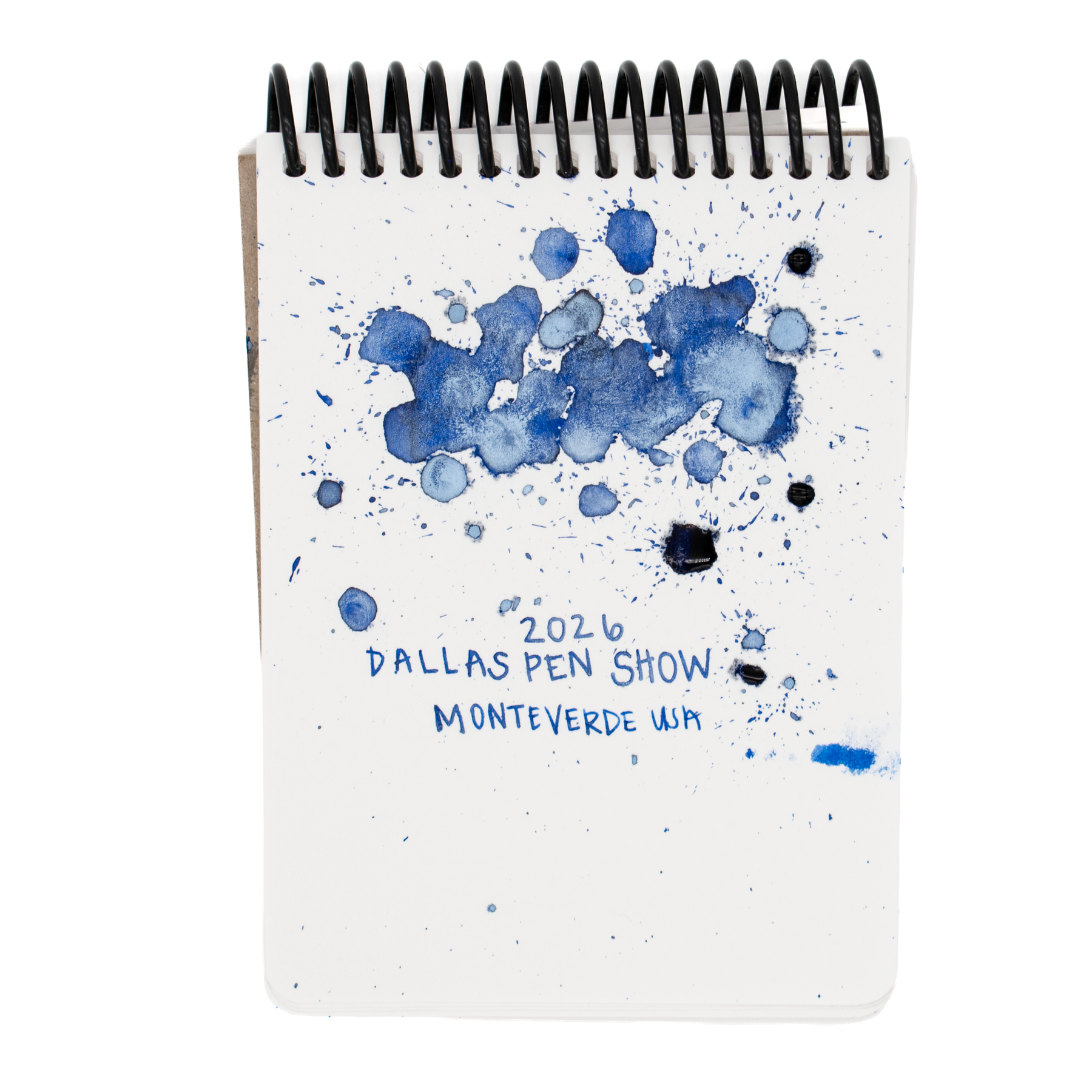 A white spiral-bound notebook featuring blue ink splatters with fine pearlescent light blue shimmer in certain spots and the text '2026 DALLAS PEN SHOW' and 'MONTVERDE USA' printed on the cover, shown against a plain white background.