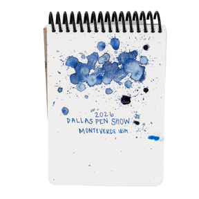 A white spiral-bound notebook featuring blue ink splatters with fine pearlescent light blue shimmer in certain spots and the text '2026 DALLAS PEN SHOW' and 'MONTVERDE USA' printed on the cover, shown against a plain white background.