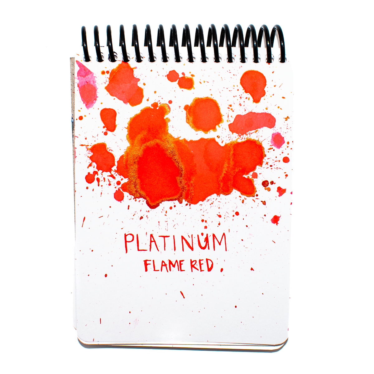Platinum's 'Flame Red' ink swatch on a white notepad with black wire bound spirals on top. There are hot pink ink splatters in the less saturated areas of the splatter droplets and a vibrant red with marbled gold and green sheen in heavily saturated areas. 