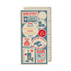 Two regular sized pencil boards stacked with cream, muted blue, red, and gold details. The illustrations showcase highlighted elements for the 2027 Traveler's Company theme 'Every Traveler's notebook is unique and that's wonderful'-- 'music lover's' 'coffee lover's' 'outdoor lover's' 'stamp lover's' are written with the illustrations. The pencil boards are on a plain white background.